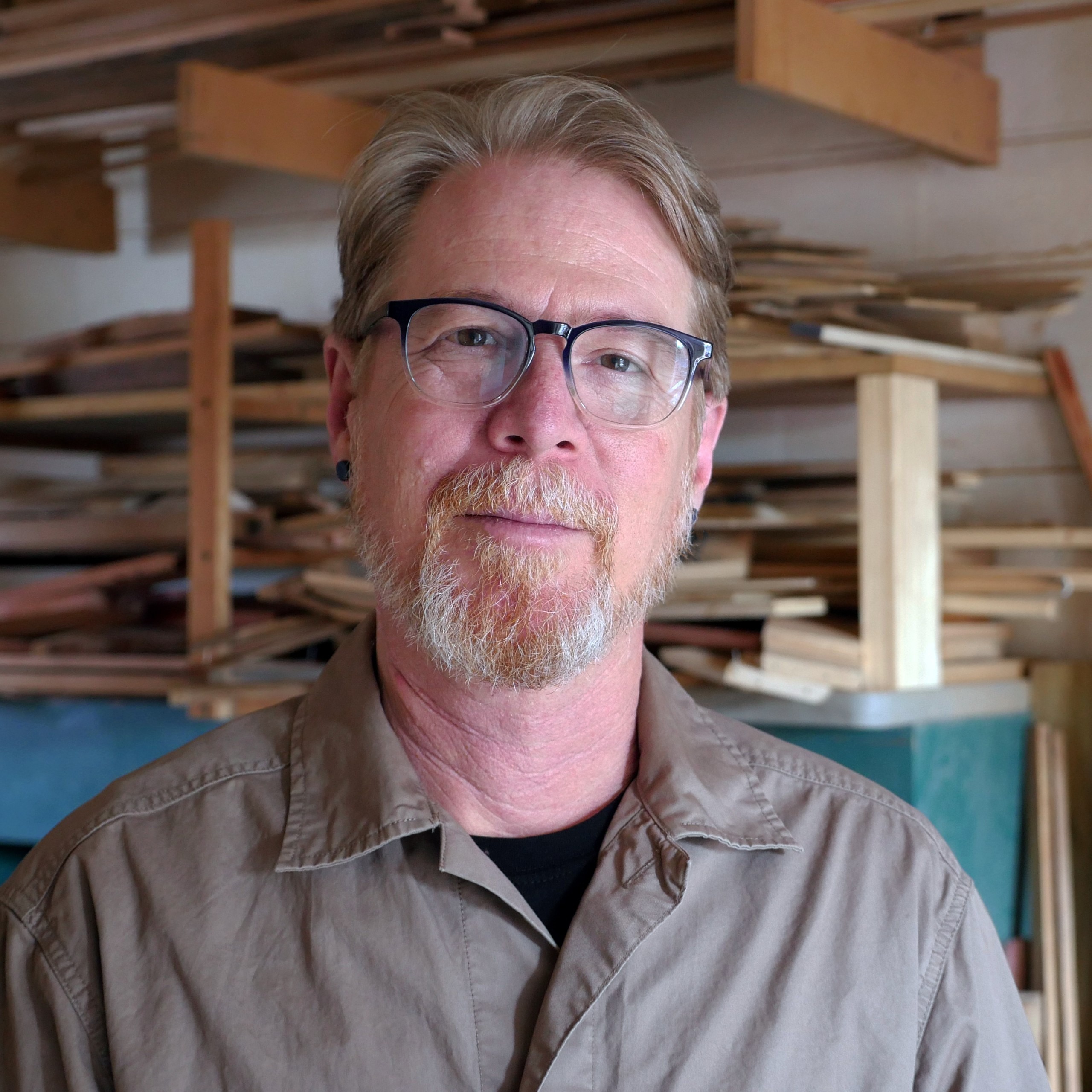 A man with glasses poses in a woodworking studio. 