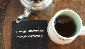 The book "The Fermi Paradox" by Courtney Clute sits flat on a table next to a cup of tea and a potted succulent.
