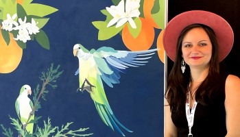 Elizabeth Barenis is shown wearing a pink hat and standing next to her painting of two monk parakeets on a navy blue background. One parakeet is perched on a cypress branch; the other is mid-flight with wings spread. Behind and overhead are oranges and orange blossoms hanging.
